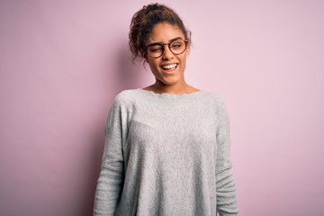 Young beautiful african american girl wearing sweater and glasses over pink background winking...