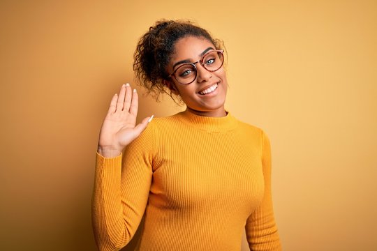 Young beautiful african american girl wearing sweater and glasses over yellow background Waiving saying hello happy and smiling, friendly welcome gesture