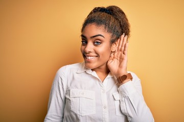 Fototapeta na wymiar Young beautiful african american girl wearing casual shirt standing over yellow background smiling with hand over ear listening an hearing to rumor or gossip. Deafness concept.