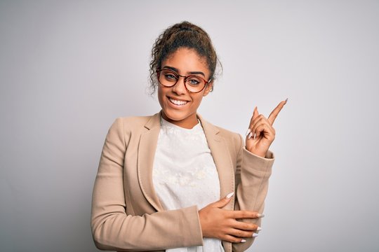 Beautiful african american businesswoman wearing jacket and glasses over white background with a big smile on face, pointing with hand and finger to the side looking at the camera.