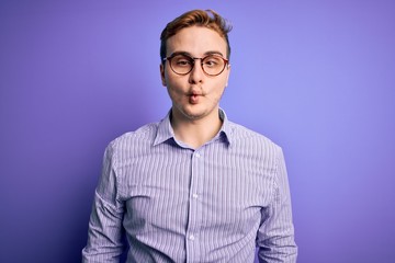 Fototapeta premium Young handsome redhead man wearing casual shirt and glasses over purple background making fish face with lips, crazy and comical gesture. Funny expression.