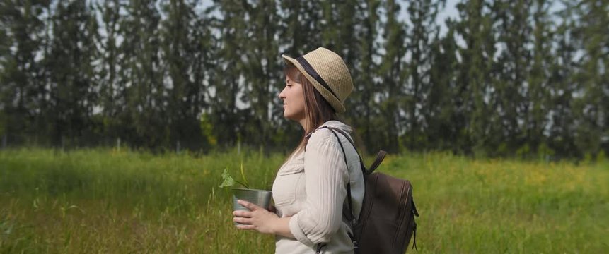 Young woman with hat, backpack and old film camera walking through the green field, carrying a pot plant. Earth day, environment, new life concept. MEDIUM SHOT, SLOW MOTION, BMPCC 4K 