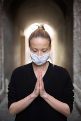 Coronavirus outbreak. Young caucasian woman wearing medical protection face mask praying over coronavirus global pandemic, for salvation of humanity, health, anxiety and depression reduction
