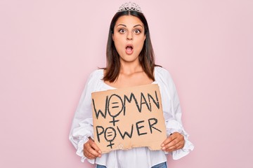 Girl wearing princess tiara asking for women rights holding banner with woman power message scared and amazed with open mouth for surprise, disbelief face