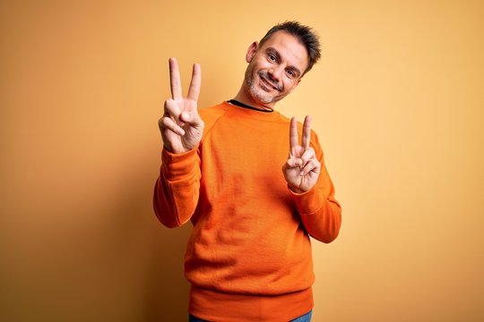 Young handsome man wearing orange casual sweater standing over isolated yellow background smiling looking to the camera showing fingers doing victory sign. Number two.