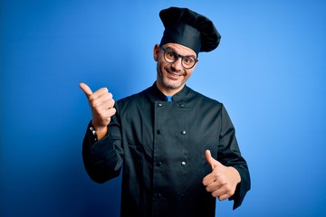 Obraz premium Young handsome chef man wearing cooker uniform and hat over isolated blue background Pointing to the back behind with hand and thumbs up, smiling confident