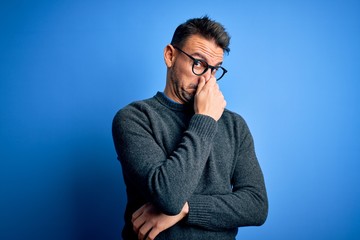 Young handsome man wearing casual sweater and glasses standing over blue background smelling something stinky and disgusting, intolerable smell, holding breath with fingers on nose. Bad smell