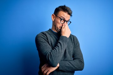 Young handsome man wearing casual sweater and glasses standing over blue background smelling...