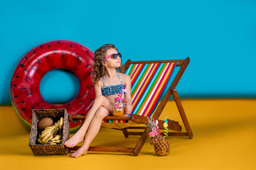 Girl wearing swimsuit, sunglasses holding jar with juice or cocktail with multicolored straws sitting in rainbow deck chair by big watermelon inflatable circle and looking away.