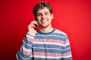 Young blond man with curly hair having conversation talking by the smartphone with a happy face standing and smiling with a confident smile showing teeth