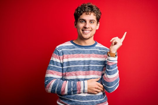 Young blond handsome man with curly hair wearing striped sweater over red background with a big smile on face, pointing with hand and finger to the side looking at the camera.