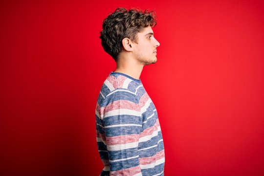 Young blond handsome man with curly hair wearing striped sweater over red background looking to side, relax profile pose with natural face with confident smile.