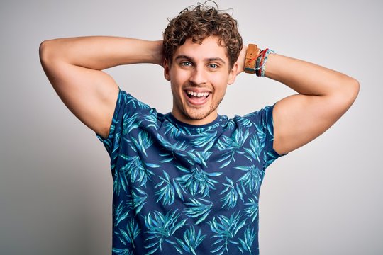 Young blond handsome man on vacation with curly hair wearing casual summer t-shirt relaxing and stretching, arms and hands behind head and neck smiling happy