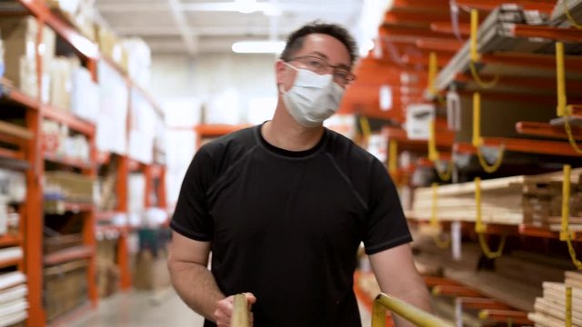 Man with face mask shopping for lumber wood in hardware store during global pandemic for home project with PPE on