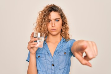 Young beautiful blonde woman with blue eyes drinking glass of water over white background pointing with finger to the camera and to you, hand sign, positive and confident gesture from the front