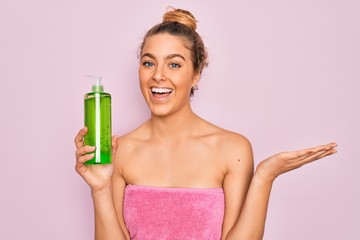 Beautiful blonde woman with blue eyes wearing towel shower after bath holding aloe vera gel very happy and excited, winner expression celebrating victory screaming with big smile and raised hands