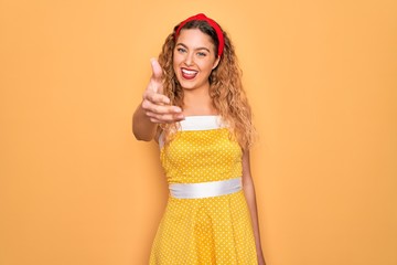 Beautiful blonde pin-up woman with blue eyes wearing diadem standing over yellow background smiling friendly offering handshake as greeting and welcoming. Successful business.