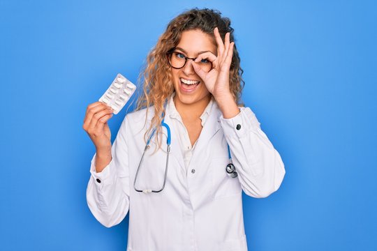 Young beautiful blonde doctor woman with blue eyes wearing stethoscope holding pills with happy face smiling doing ok sign with hand on eye looking through fingers