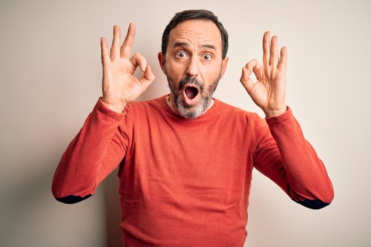 Middle age hoary man wearing casual orange sweater standing over isolated white background looking surprised and shocked doing ok approval symbol with fingers. Crazy expression