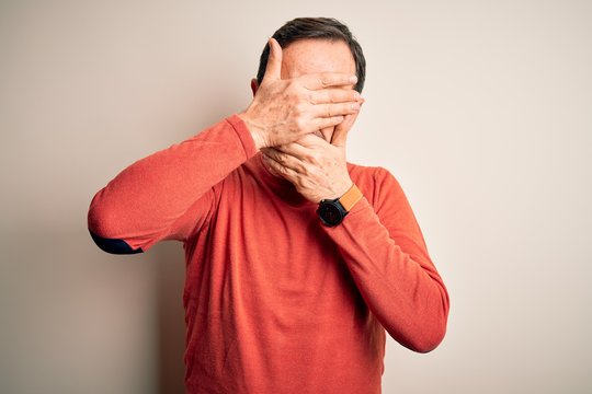 Middle age hoary man wearing casual orange sweater standing over isolated white background Covering eyes and mouth with hands, surprised and shocked. Hiding emotion