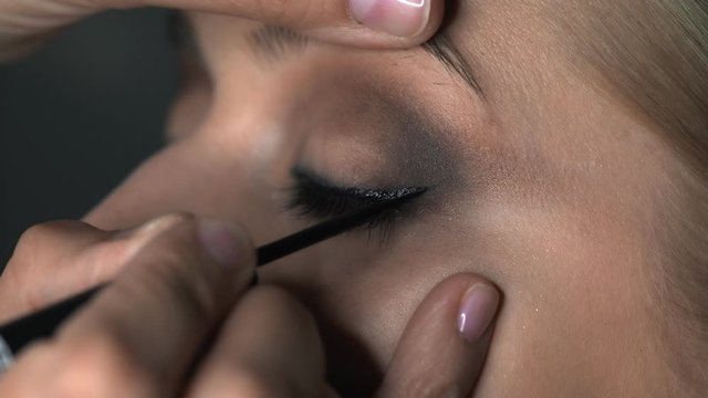 Close up of makeup artist making professional make-up for young woman in beauty studio. Make up Artist draws arrows over eye, eyeliner