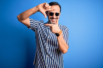 Middle age hoary man wearing striped shirt and sunglasses over isolated blue background smiling making frame with hands and fingers with happy face. Creativity and photography concept.