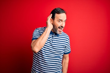 Middle age hoary man wearing casual striped polo standing over isolated red background smiling with hand over ear listening an hearing to rumor or gossip. Deafness concept.