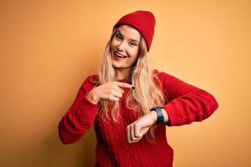 Young beautiful blonde woman wearing casual sweater and wool cap over white background In hurry pointing to watch time, impatience, upset and angry for deadline delay