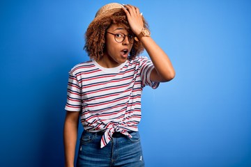 African American woman with curly hair on vacation wearing summer hat and striped t-shirt surprised...