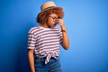 African American woman with curly hair on vacation wearing summer hat and striped t-shirt tired rubbing nose and eyes feeling fatigue and headache. Stress and frustration concept.