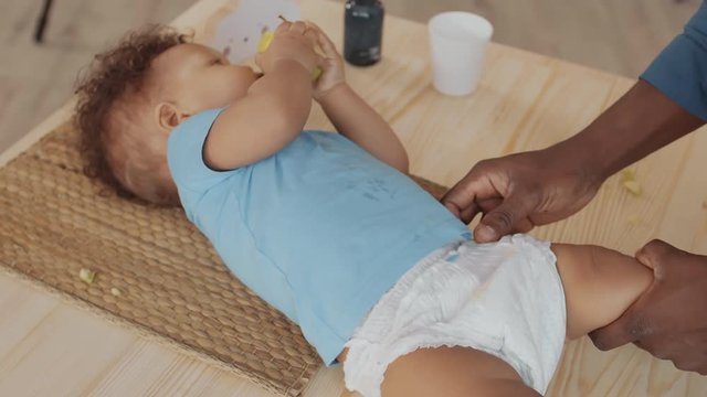 High angle view of male hands putting new disposable diaper on little African boy lying on wooden table and eating apple