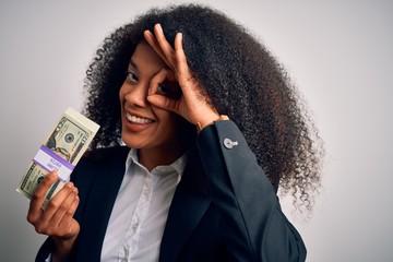 Young african american business woman with afro hair holding a bunch of 20 dollars banknotes with happy face smiling doing ok sign with hand on eye looking through fingers