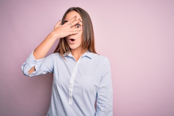 Young beautiful businesswoman wearing elegant shirt standing over isolated pink background peeking in shock covering face and eyes with hand, looking through fingers with embarrassed expression.