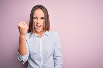Young beautiful businesswoman wearing elegant shirt standing over isolated pink background angry and mad raising fist frustrated and furious while shouting with anger. Rage and aggressive concept.