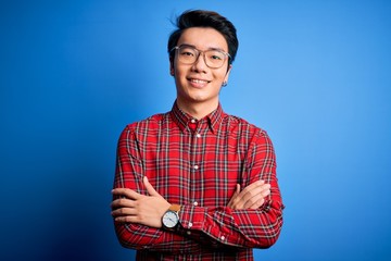 Young handsome chinese man wearing casual shirt and glasses over blue background happy face smiling...