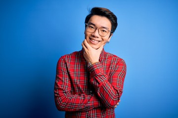 Fototapeta premium Young handsome chinese man wearing casual shirt and glasses over blue background looking confident at the camera smiling with crossed arms and hand raised on chin. Thinking positive.