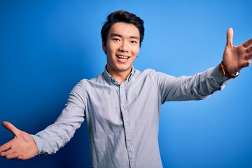 Young handsome chinese man wearing casual shirt standing over isolated blue background looking at the camera smiling with open arms for hug. Cheerful expression embracing happiness.