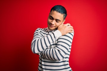 Young brazilian man wearing casual striped t-shirt standing over isolated red background Hugging...