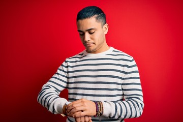 Young brazilian man wearing casual striped t-shirt standing over isolated red background Checking the time on wrist watch, relaxed and confident