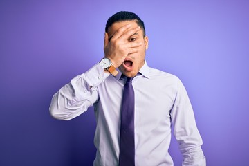 Young brazilian businessman wearing elegant tie standing over isolated purple background peeking in shock covering face and eyes with hand, looking through fingers with embarrassed expression.