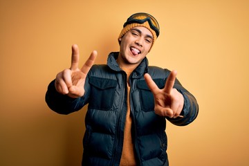 Young brazilian skier man wearing snow sportswear and ski goggles over yellow background smiling with tongue out showing fingers of both hands doing victory sign. Number two.