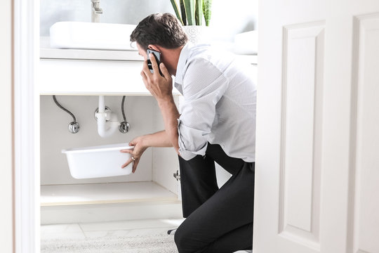 Man Calling Plumber In Front Of Water Leaking From Sink Pipe