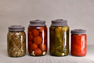 Four glass jars in a row with textile that against rust, with fermented cucumbers, tomatoes, mushrooms and bell peppers.