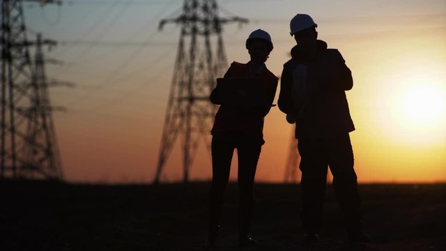 silhouette, engineers in protective helmets with drawings and a laptop in their hands discuss development of electric lines background of power line toppers and sunset