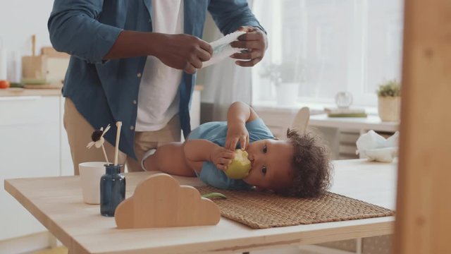 Lockdown of unrecognizable dad changing disposable diaper on African curly-haired toddler lying on kitchen table and eating fresh green apple