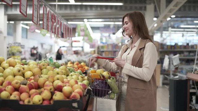 lifestyle, happy young woman shopper with food basket in hands picks fresh apple fruit in supermarket, commerce