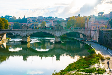 Fototapeta premium Rome, Italy. Ponte Vittorio Emanuele II bridge spans River Tiber in the Italian city. Public/ private transportation drive across, pedestrians walk along & on street level below. Old buildings behind.