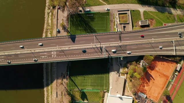 Ascending aerial drone top view of a highway bridge with a lot of traffic. The bridge is running over a strangely placed soccer/football field. Abstract concept of modern transportation & engineering.