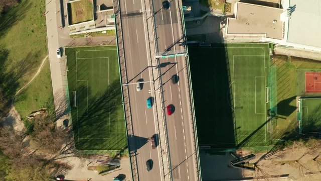 Ascending aerial drone top view of a highway bridge with a lot of traffic. The bridge is running over a strangely placed soccer/football field. Abstract concept of modern transportation & architecture