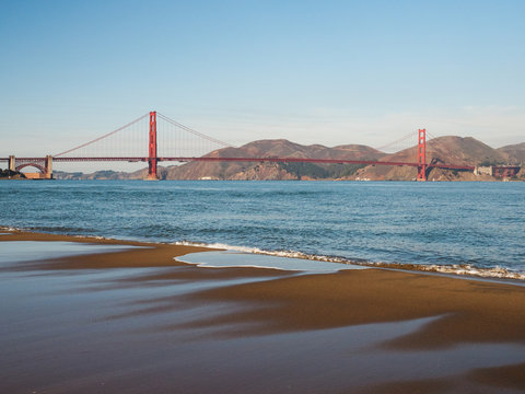The famous Golden Gate Bridge shot at midday. The image of the bridge is reflected in the water on the sand. 
